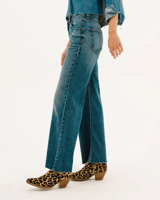 A person wearing ASKK NY’s Mercer Jean in Gunnison, a denim shirt, and leopard-print heeled ankle boots stands against a plain white background.