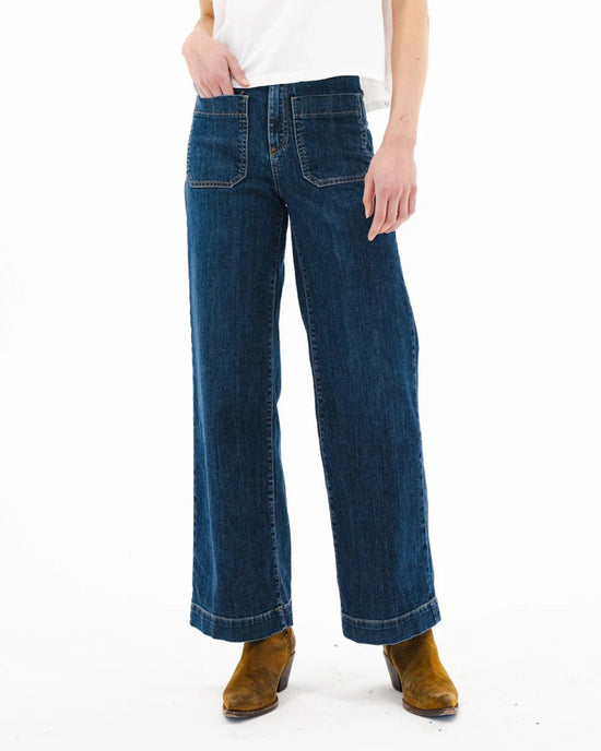 A person wearing a white shirt, ASKK NY Sailor in Dark Stone high rise wide leg jeans with front patch pockets, and brown suede ankle boots, stands against a plain white background.