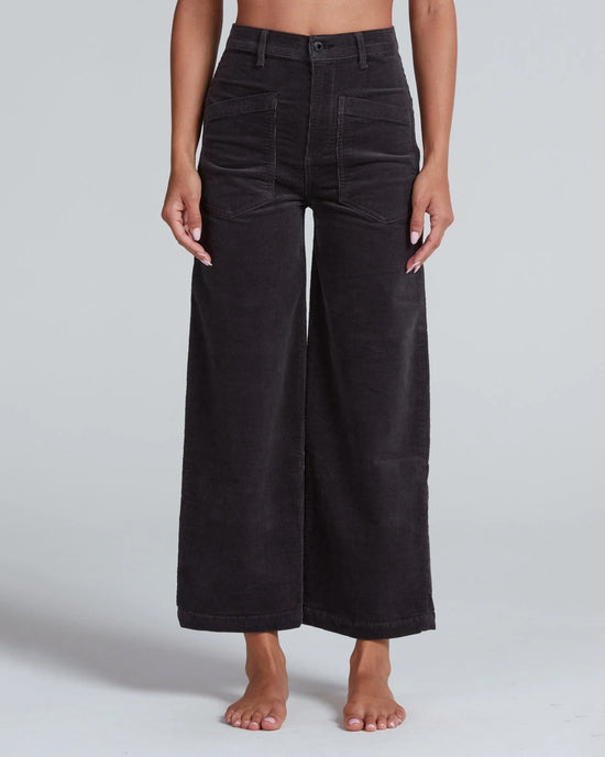 A person stands barefoot against a plain light gray background wearing ASKK NY’s Virginia Corduroy in Charcoal—high-rise, wide-leg pants featuring front patch pockets, visible stitching, and a button fly.