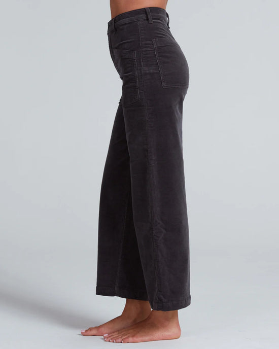 A person stands barefoot in profile against a plain light gray background, wearing ASKK NY’s Virginia Corduroy in Charcoal—high-rise, wide-leg pants with front patch pockets and a cropped length.