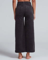 A person stands barefoot, shown from the back, wearing ASKK NY Virginia Corduroy wide-leg, high-rise pants in Charcoal with visible back pockets and seam details. The cropped pants contrast against a plain light gray background.