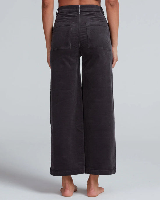 A person stands barefoot, shown from the back, wearing ASKK NY Virginia Corduroy wide-leg, high-rise pants in Charcoal with visible back pockets and seam details. The cropped pants contrast against a plain light gray background.