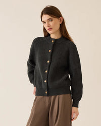 A woman with long brown hair wears the Demylee Audrey Cotton Cardigan in Dark Charcoal, paired with loose brown pants, standing against a plain light background and looking slightly to the side with a neutral expression.