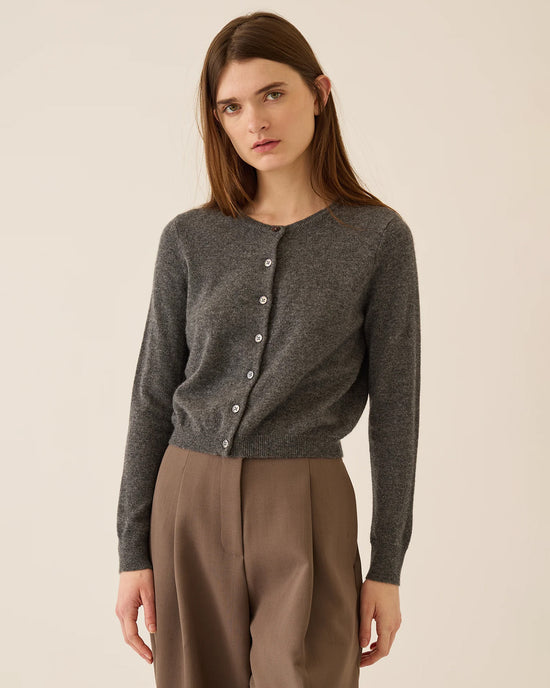 A woman with straight brown hair wears the Demylee Ava Cashmere Cardigan in Dark Heather Grey and high-waisted brown trousers, standing against a plain beige background.