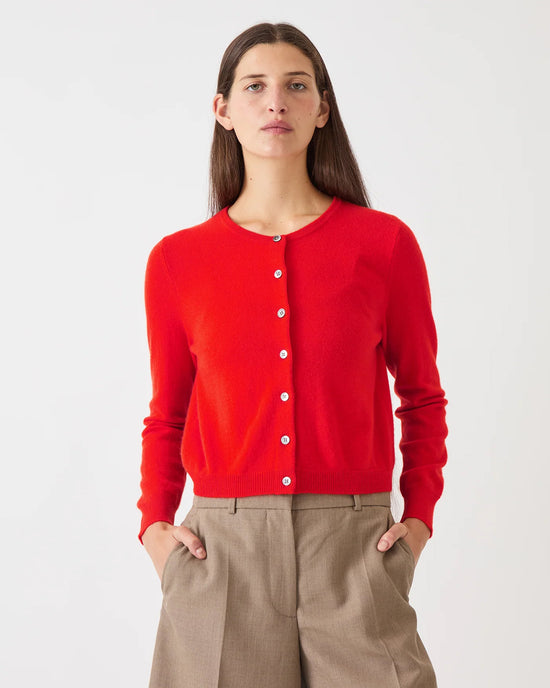 A woman with long brown hair wears the Demylee Ava Cashmere Cardigan in Scarlet and high-waisted beige trousers, standing with her hands in her pockets against a plain white background.