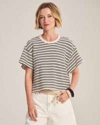 A woman with short blonde hair wears the Brissa Short Sleeve Crew Neck Top in Natural/Navy by Velvet by Graham & Spencer and white pants. She stands against a plain background, smiling slightly with one hand in her pocket.