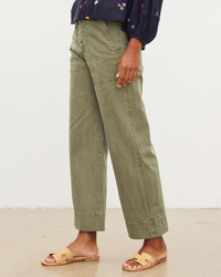 A person standing in Mya Pants in Axe by Velvet by Graham & Spencer and yellow sandals, with a dark slim top featuring small floral embroidery details.