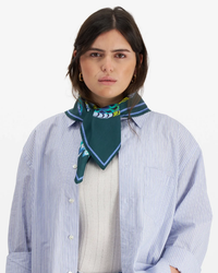 A woman wearing a striped blue and white shirt over a cream sweater, accessorized with a luxury Inoui Editions Square / Carre 65 Hulule in Emerald scarf.