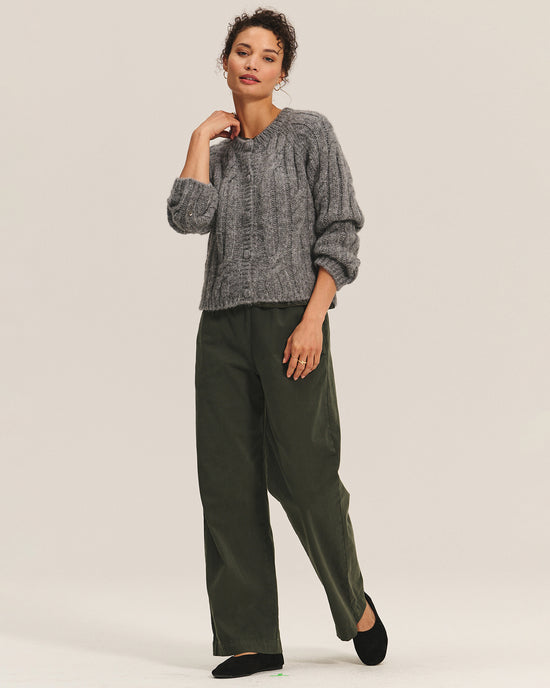 A woman stands against a plain background wearing the Velvet by Graham & Spencer Allison Long Sleeve Crew Cardi in Heather Grey, paired with loose olive green pants and black shoes. She has curly hair tied back, posing with one hand on her neck and the other in her pocket.