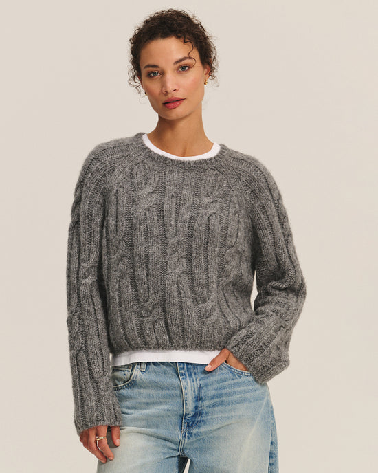 A woman with short curly hair wears the Marina Long Sleeve Crew Top in Heather Grey by Velvet by Graham & Spencer over a white shirt and light blue jeans, standing confidently against a plain background.