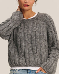 A woman wears the Marina Long Sleeve Crew Top in Heather Grey by Velvet by Graham & Spencer over a white shirt with blue jeans. One arm is raised near her face; her lips are visible but eyes are out of frame.