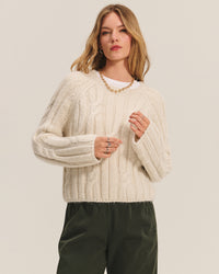 A woman with long, light brown hair wears the Marina Long Sleeve Crew Top in Ivory by Velvet by Graham & Spencer, paired with dark green pants and a gold chain necklace, standing against a plain light background.