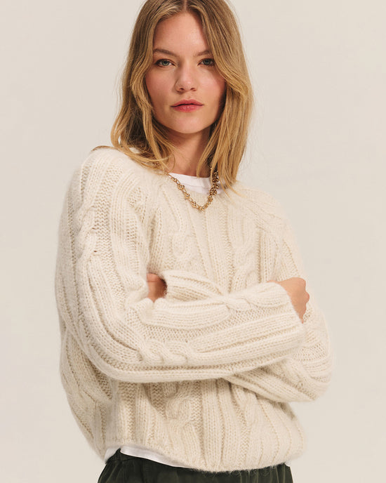 A woman with long blonde hair wears the Velvet by Graham & Spencer Marina Long Sleeve Crew Top in Ivory over a white shirt and gold chain necklace. She stands with arms crossed, looking confidently at the camera against a plain background.