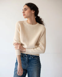 A woman with curly dark hair, dressed in a Demylee Chelsea Washed Cotton Sweater in Off White and blue jeans, stands against a plain light background, looking to the side with one arm gently resting across her body.