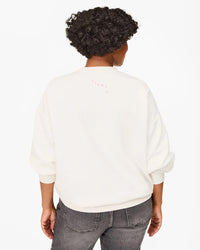 A person with short curly hair is seen from behind in a Clare V. "Iconique" Oversized Sweatshirt in Oyster White w/ Peony, made of 100% cotton fleece, and faded black jeans, set against a plain white background.