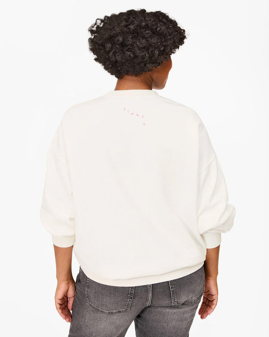 A person with short curly hair is seen from behind in a Clare V. "Iconique" Oversized Sweatshirt in Oyster White w/ Peony, made of 100% cotton fleece, and faded black jeans, set against a plain white background.