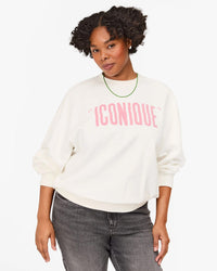 A woman with curly hair wears the Clare V. "Iconique" Oversized Sweatshirt in Oyster White with Peony lettering, styled with dark jeans and a green necklace, standing against a plain white background. Made from 100% cotton fleece.