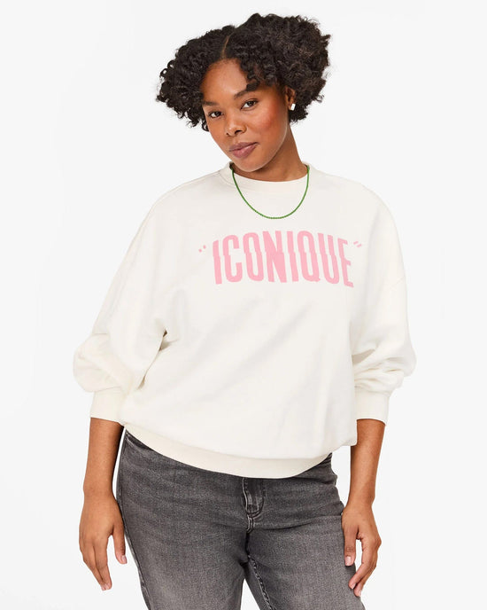 A woman with curly hair wears the Clare V. "Iconique" Oversized Sweatshirt in Oyster White with Peony lettering, styled with dark jeans and a green necklace, standing against a plain white background. Made from 100% cotton fleece.
