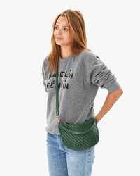 A woman with long brown hair wears a grey sweatshirt that says Masculin Féminin, light blue jeans, and carries the Clare V. OLD-Woven Zig-Zag Petit Moyen Messenger in Evergreen, standing against a plain white background.