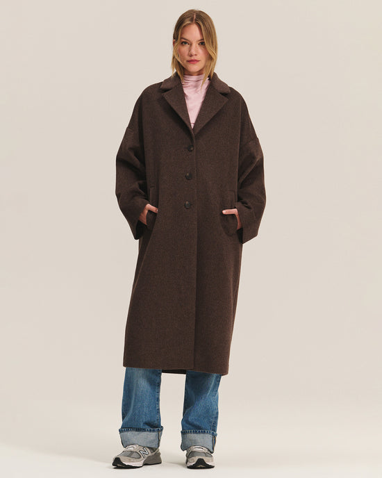 A woman faces forward with her hands in the pockets of the Velvet by Graham & Spencer Lafayette Long Coat in Mocha, styled over a light pink turtleneck, blue jeans, and gray sneakers against a plain light background.