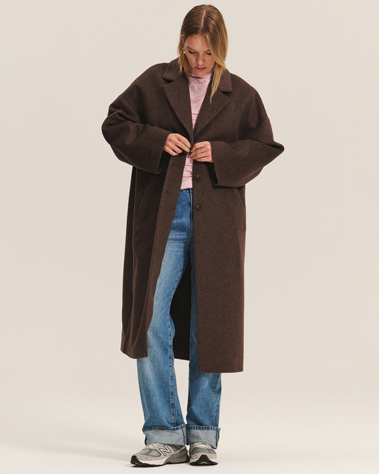 A person wearing the Lafayette Long Coat in Mocha by Velvet by Graham & Spencer over a pink shirt, blue jeans, and gray sneakers fastens a button on the coat while looking down against a plain background.