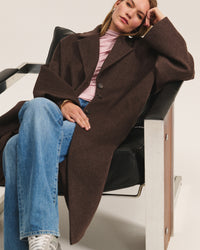 A woman sits in a black and metal chair, wearing the Lafayette Long Coat in Mocha by Velvet by Graham & Spencer over a pink turtleneck, blue jeans, and gray sneakers. She rests her head on her hand, looking relaxed.