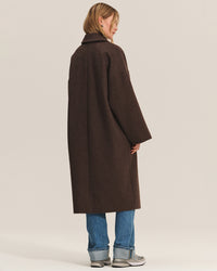 A person with short blonde hair faces away, wearing the Lafayette Long Coat in Mocha by Velvet by Graham & Spencer, paired with blue jeans and gray sneakers against a plain light background.