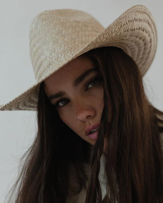 A woman with long brown hair wearing a Gigi Pip Codi Western in Natural straw hat, gazing intently at the camera with a slight pout.