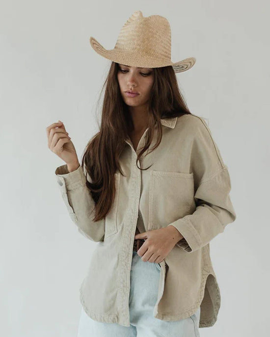 A woman in a Gigi Pip Codi Western straw hat with a pinched fedora crown and oversized beige shirt, with one hand on her hip, looks down thoughtfully.