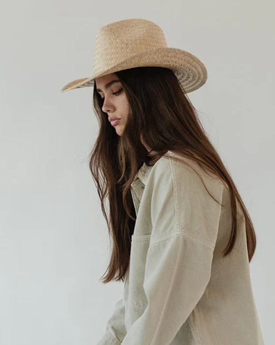 Woman in a Gigi Pip Codi Western in Natural straw hat with a pinched fedora crown and beige shirt, looking downwards with her long hair cascading over her shoulders, against a plain background.