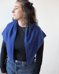 A woman with long, wavy brown hair, wearing a black sweater and blue jeans, stands facing left against a plain, light background. She is wearing the COLORI Large Triangle Bandana in Neptune.