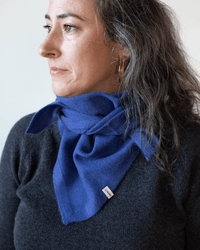 A woman with wavy dark hair wears a dark sweater and the COLORI Large Triangle Bandana in Neptune, tied around her neck. She looks to the left, showing a small tag on the lightweight bandana.
