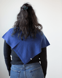 A person with long, wavy dark hair, tied with a green clip, wears a COLORI Large Triangle Bandana in Neptune over a black sweater and blue jeans. The lightweight bandana enhances the outfit with casual elegance against a plain light background.