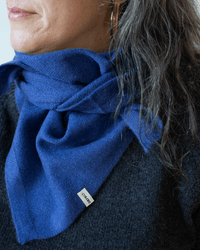 A close-up of a woman with gray hair wearing a COLORI Large Triangle Bandana in Neptune, wrapped around her neck and featuring a small white KOMODE tag. She is dressed in a dark sweater.