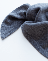 A close-up of the COLORI Small Triangle Bandana in Kensington, crafted from soft, dark gray 100% cashmere knit, arranged to resemble bunny ears on a white background.