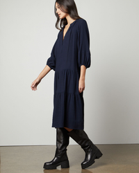Woman standing in a navy blue Velvet by Graham & Spencer Dixon Puff Slv Tier Dress paired with black knee-high boots.