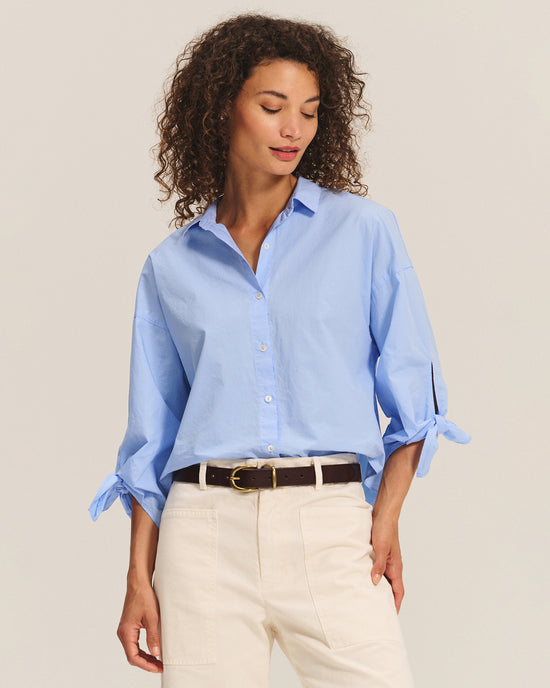 A woman with curly hair wears the Velvet by Graham & Spencer Arlette Knot Sleeve Button Up in Chill and off-white pants, accessorized with a black belt. She looks down slightly, standing against a plain light background.