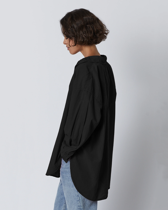 A person standing side-on wearing a Velvet by Graham & Spencer Redondo L/S Button Up in Black in an oversized fit and denim jeans against a neutral background.