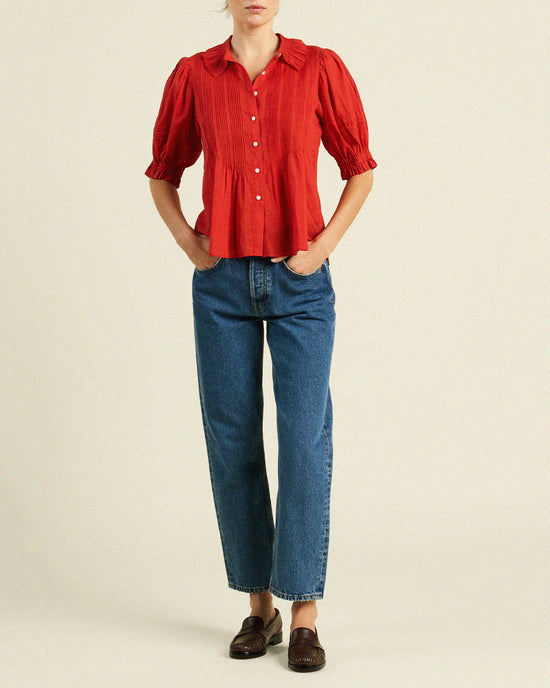 A person in a Jess Blouse in Crimson Check by Trovata Birds of Paradis, paired with blue straight-leg jeans and dark brown loafers, stands with hands in pockets against a plain background.