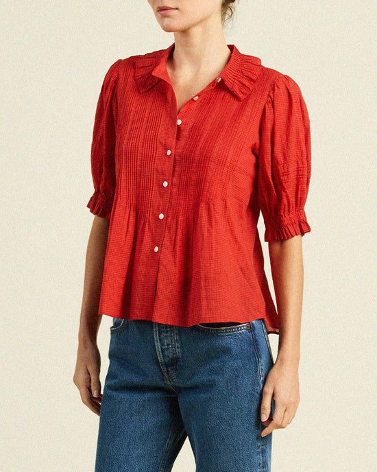 A woman wears a Trovata Birds of Paradis Jess Blouse in Crimson Check with blue jeans, standing against a plain light background.