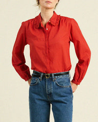 Wearing the Trovata Birds of Paradis Kinsley Blouse in Crimson Check, a long-sleeved 100% cotton top with a ruffled collar, tucked into high-waisted blue jeans with a black belt, the person stands against a plain light background.