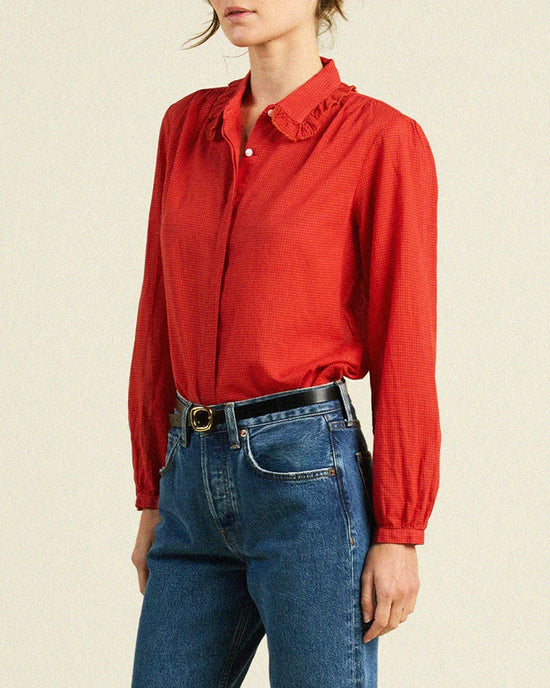 A woman in the Trovata Birds of Paradis Kinsley Blouse in Crimson Check—a long-sleeved red cotton top with a ruffled collar—paired with blue jeans stands before a plain beige background, her face partially turned out of frame.