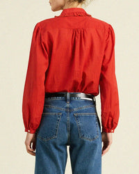A person wearing the Trovata Birds of Paradis Kinsley Blouse in Crimson Check, a 100% cotton top with a ruffled collar, paired with blue high-waisted jeans, seen from behind against a plain light background.