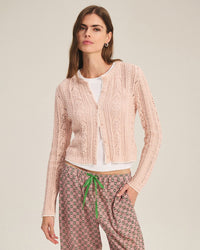 A woman with long brown hair wears the Velvet by Graham & Spencer Daisy Long Sleeve Crew Cardi in Blush over a white top and patterned pants with a green drawstring, standing against a plain background.