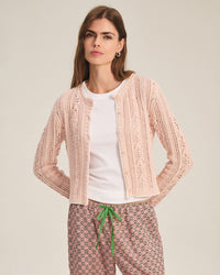 A woman with long brown hair wears the Daisy Long Sleeve Crew Cardi in Blush by Velvet by Graham & Spencer over a white T-shirt and patterned pants with a green drawstring, standing with her hands behind her back against a plain background.