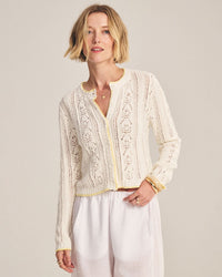 A woman with short blonde hair wears the Daisy Long Sleeve Crew Cardi in Ivory by Velvet by Graham & Spencer, a cream-colored, cropped cardigan with white buttons and a delicate pattern. She pairs it with loose white pants and smiles softly against a light background.