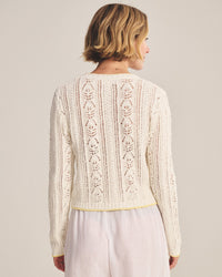 A woman with short, wavy blonde hair is seen from behind wearing the Daisy Long Sleeve Crew Cardi in Ivory by Velvet by Graham & Spencer and light-colored, loose-fitting pants. The background is plain and neutral.