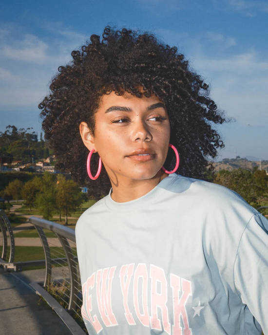 Woman with curly hair wearing large Barbie wood hoop earrings and a "new york" sweatshirt outdoors.