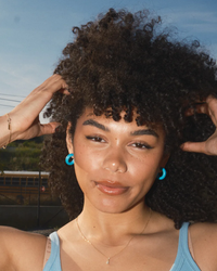 Woman with curly hair smiling at the camera, wearing B&L's C Hoop in Mini in Ken earrings with 14k gold filled posts and a light blue top.