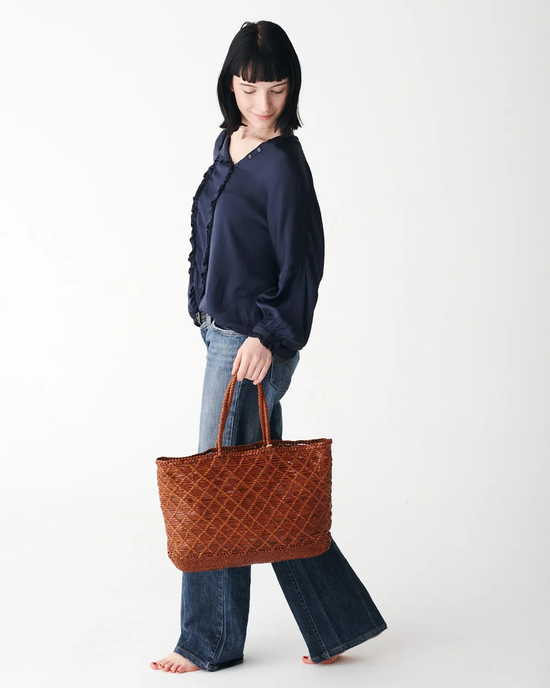 Woman with a braided hairstyle posing sideways, holding a large Dragon Diffusion EW Corso Bag in Tan with a crosshatch design, wearing a navy blouse and flared jeans.